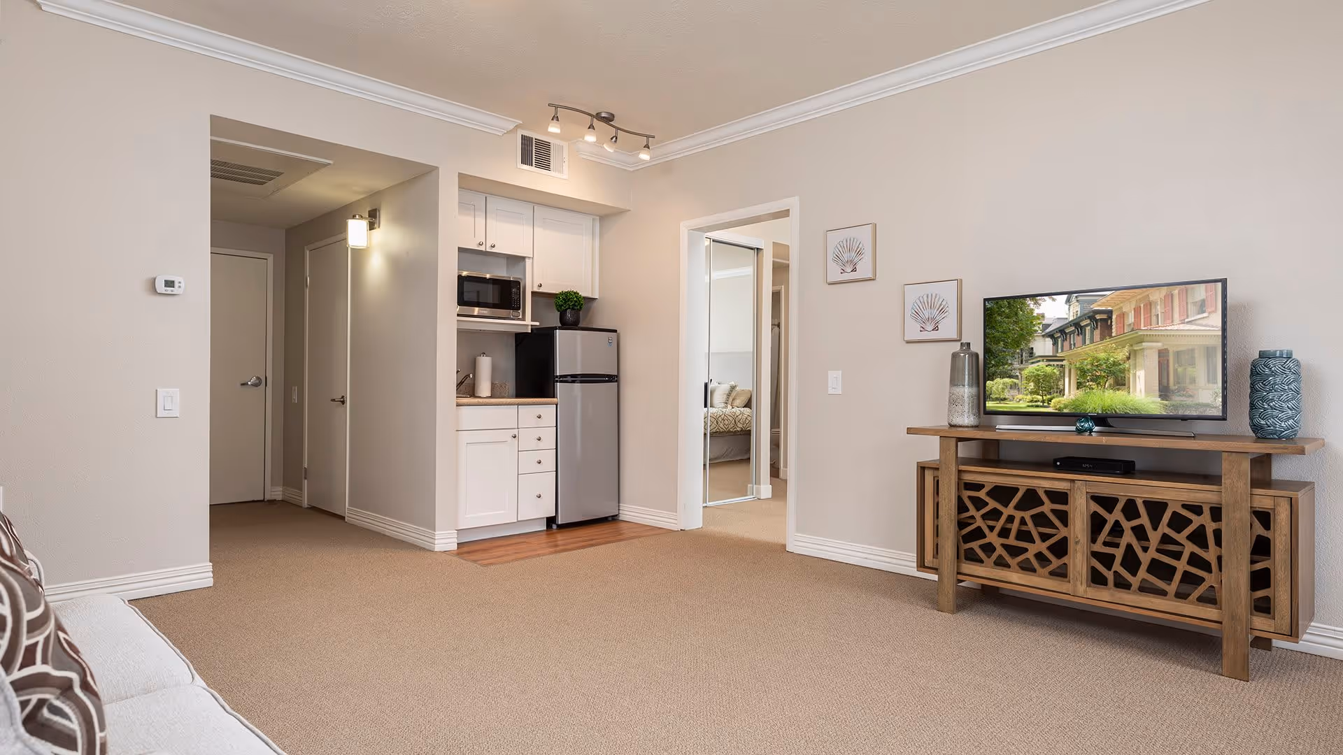 A cozy living room area with beige walls and carpet, featuring a wooden TV stand with decorative cutouts, a flat-screen TV displaying a neighborhood scene, and a small kitchenette with white cabinets, a microwave, and a mini refrigerator. A doorway leads to a bedroom with a mirrored closet door.