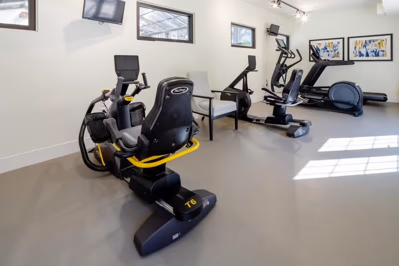 A fitness room with exercise equipment including a recumbent bike, an elliptical machine, and a treadmill. The room has light-colored walls, several small windows near the ceiling, a wall-mounted TV, and framed artwork on the far wall. There is also a gray armchair positioned between the exercise machines.