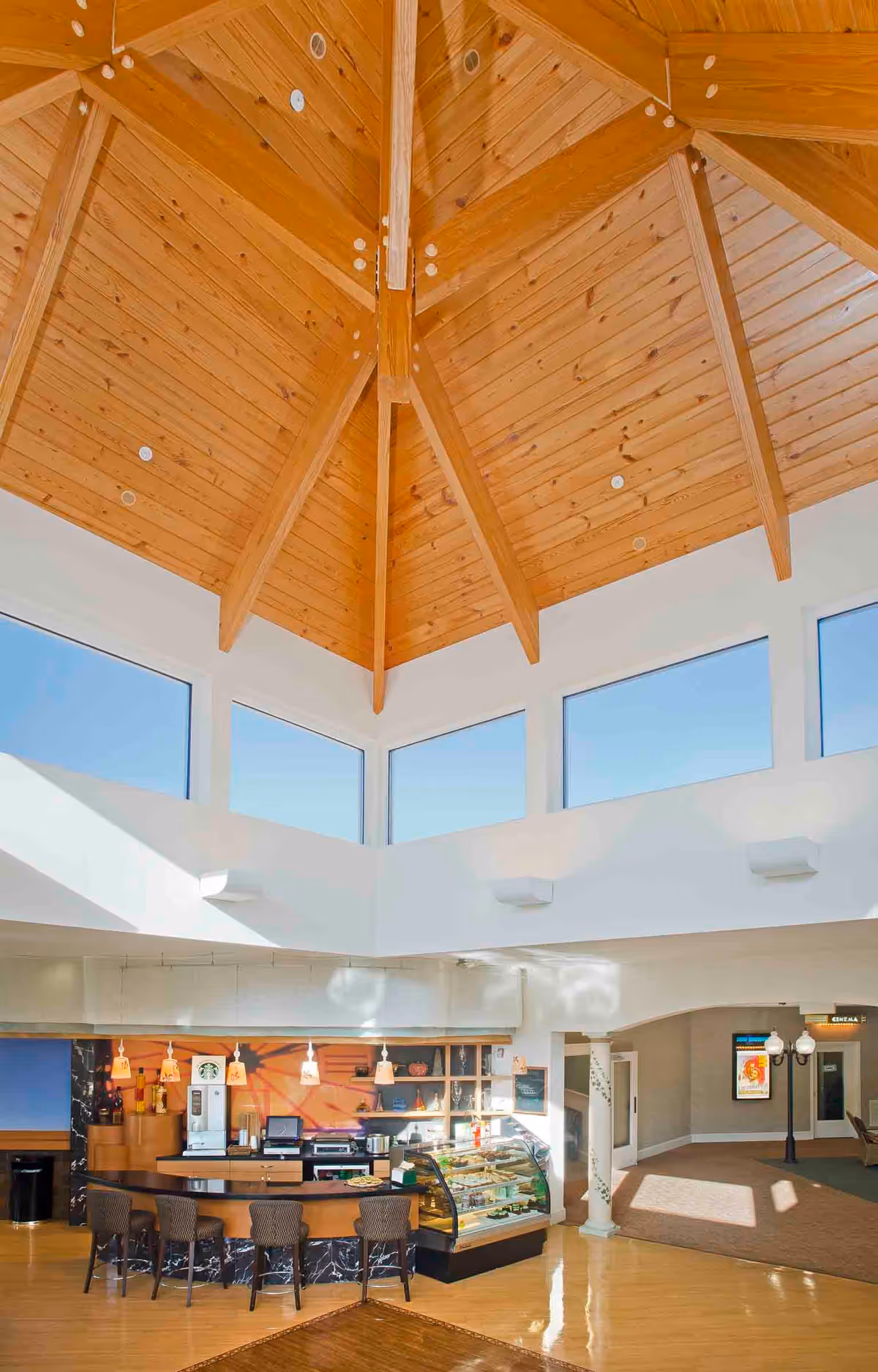 Interior view of a senior living facility cafe area with a high wooden vaulted ceiling and large windows near the ceiling. The cafe counter has several chairs, a display case with food items, and a coffee machine. The floor is wooden, and there is an open hallway leading to other parts of the facility.