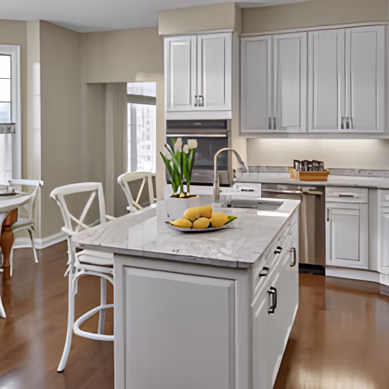 A modern kitchen with white cabinetry and a marble countertop island. The island has a sink and a plate with lemons on it. There are white chairs around the island and a dining table with chairs in the background. The kitchen has stainless steel appliances and wooden flooring.