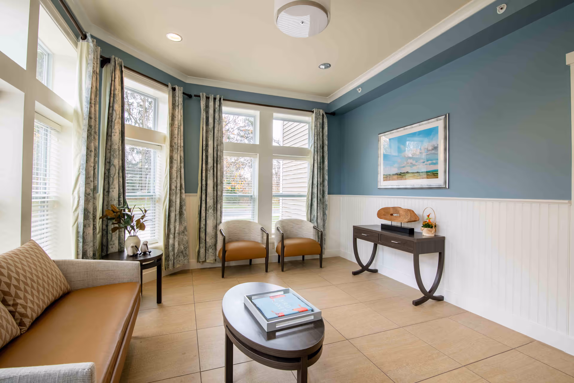 Sunlit sitting room with a sofa, two chairs, coffee table and console table beneath large windows with curtains.