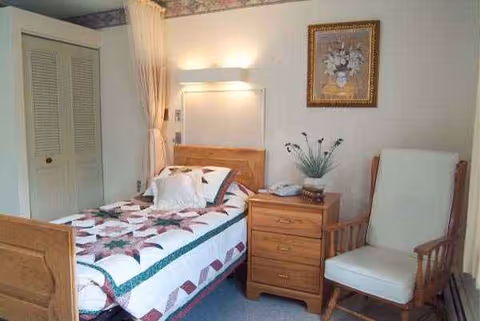 A cozy bedroom in a senior care facility with a quilted bed, dresser, and chair.