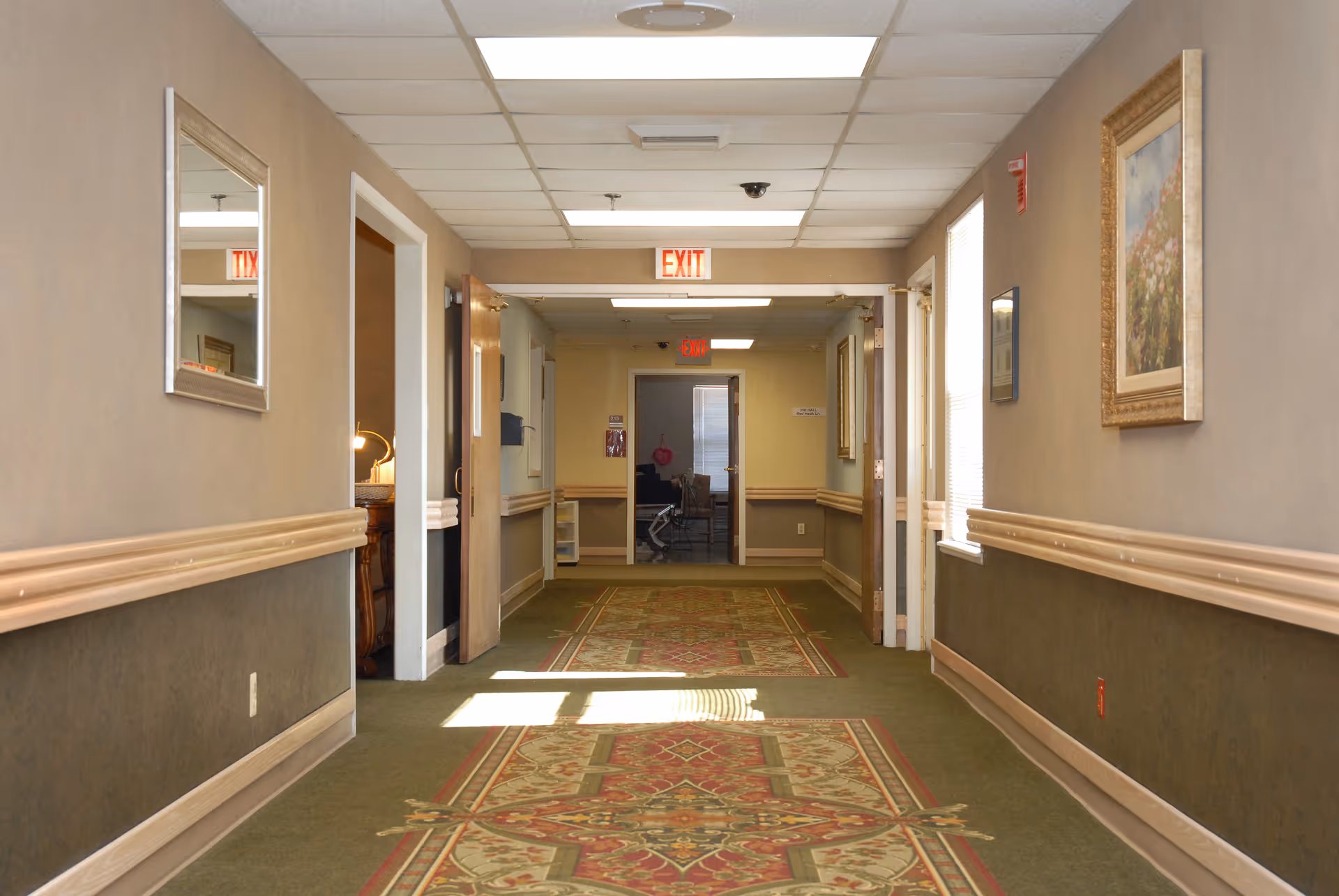 A well-lit hallway in a senior living facility with patterned carpet, beige walls with wooden chair rails, framed artwork, and an exit sign at the end. Doors and windows line the hallway, and a mirror is mounted on the left wall.