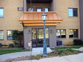 Entrance of a multi-story brick building with a covered doorway, glass double doors, a lamp post, and surrounding shrubs and sidewalk.