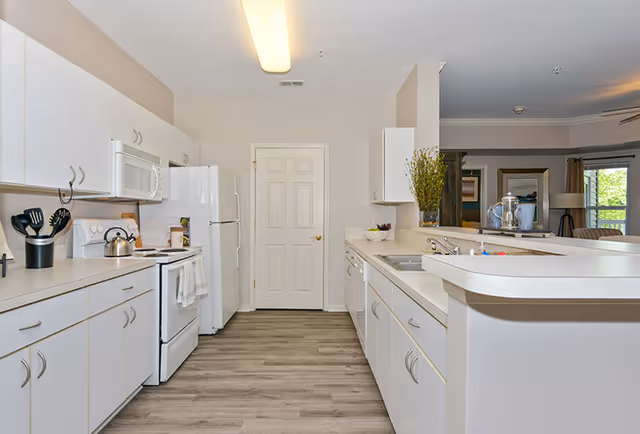 A clean, modern kitchen with white cabinets and appliances including a refrigerator, stove, microwave, and dishwasher. The kitchen has light-colored countertops and wood-style flooring. There is a door at the end of the kitchen and an open view into a living area with windows and furniture.