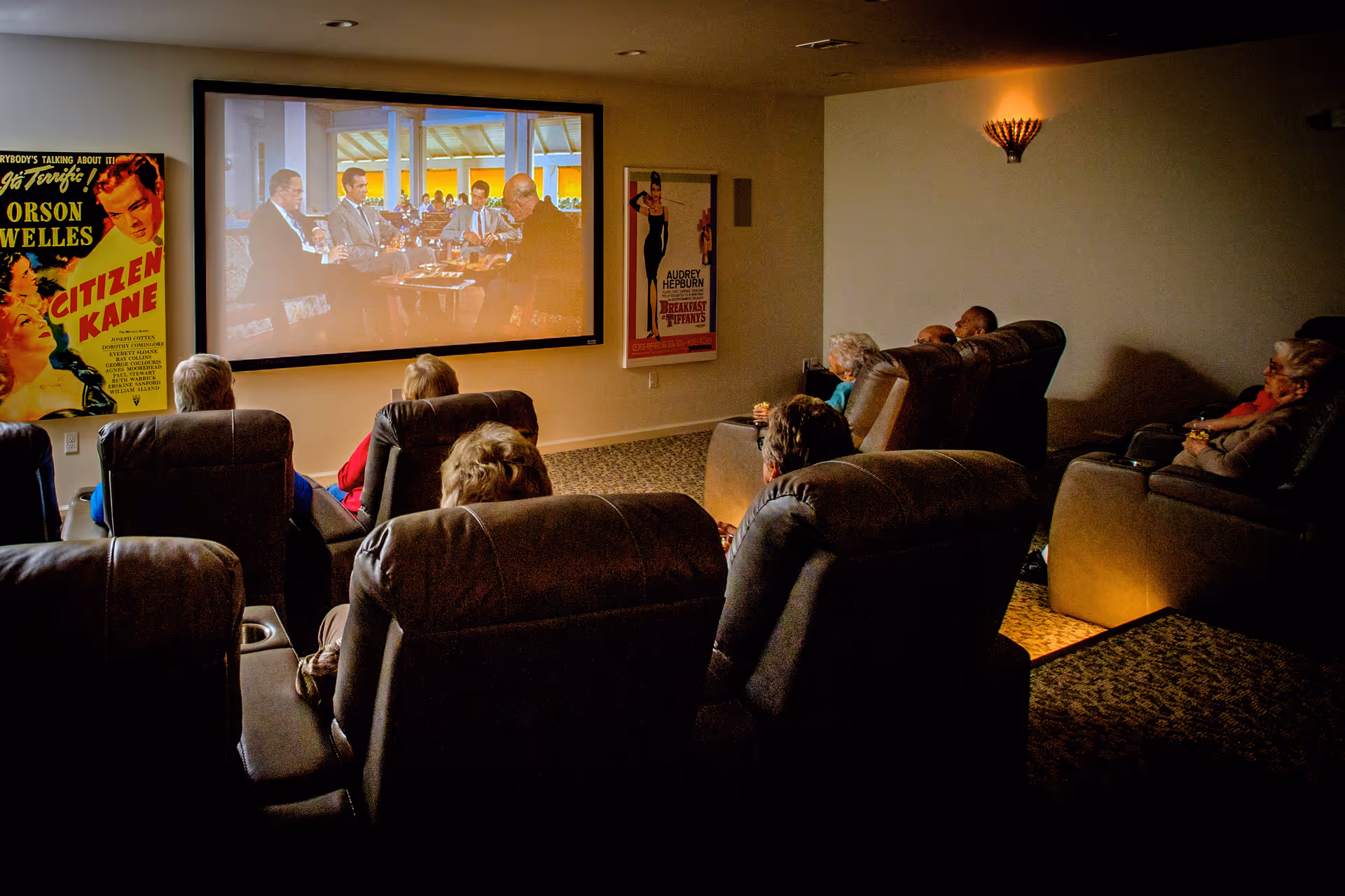 A group of elderly people seated in comfortable recliner chairs watching a movie projected on a large screen in a dimly lit room. Movie posters for Citizen Kane and Breakfast at Tiffany's are displayed on the walls.