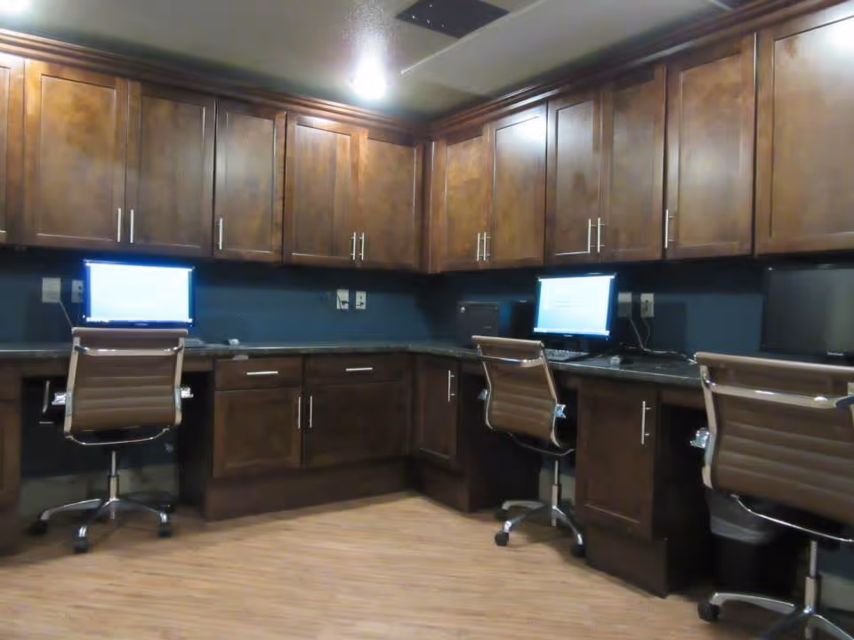 Interior office area with three brown swivel chairs in front of a long counter with three computer monitors. The counter is surrounded by wooden cabinets above and below, and the floor has a wood finish.