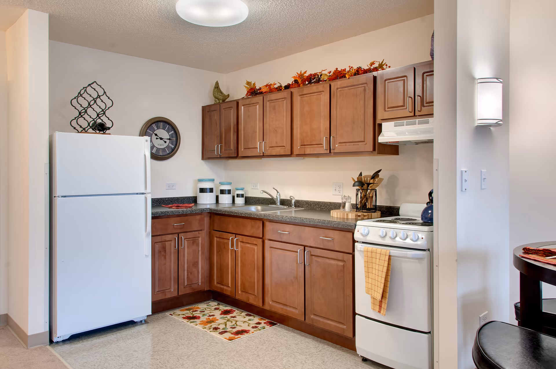 A compact kitchen with wooden cabinets, a white refrigerator, a white stove with a yellow towel hanging on the handle, a countertop with a sink, and decorative fall-themed garland on top of the cabinets. There is a wall clock and a wine rack on the wall, and a small floral rug on the floor.