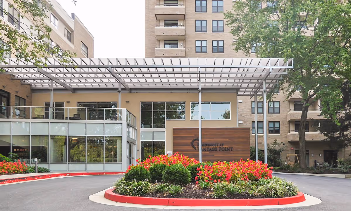 Exterior view of the Residences at Vantage Point CCRC building entrance with a covered driveway, landscaped flower bed with red and green plants, and multi-story residential building in the background.