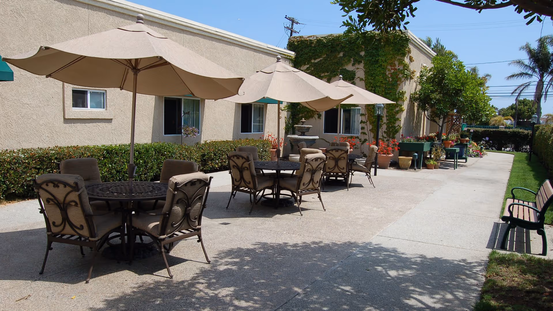 Outdoor patio area with two round tables, each surrounded by four cushioned chairs and shaded by large beige umbrellas. The patio is adjacent to a beige building with windows and is decorated with potted plants and greenery. There is a bench on the right side along a grassy area, and trees and shrubs line the walkway.