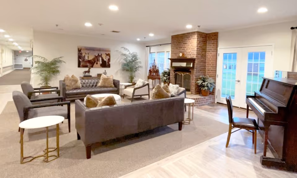 A cozy senior living common area with multiple gray sofas and armchairs arranged around small white tables. There is a brick fireplace with plants on either side, a piano with a chair, and large windows with glass doors letting in natural light. The walls are decorated with a painting of horses and some decorative items.