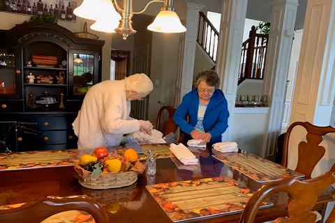 Two elderly women folding white towels at a wooden dining table with autumn-themed placemats and a basket of artificial fruit in a cozy, well-lit dining room with a black cabinet and staircase in the background.