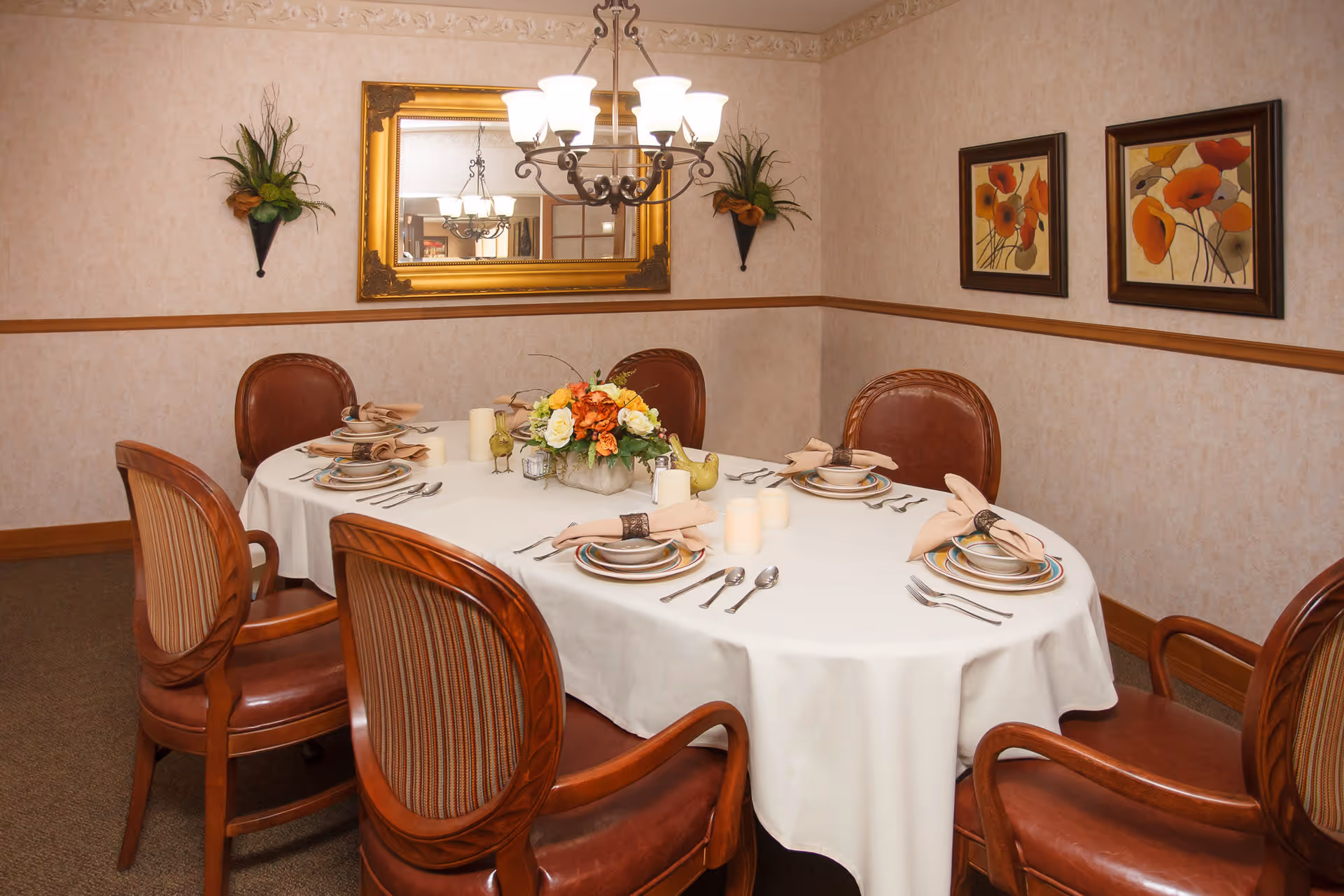 A dining room with an oval table covered with a white tablecloth, set for six people with plates, bowls, utensils, and napkins. The table has a floral centerpiece and decorative candles. The room features a chandelier, a large gold-framed mirror, two framed floral paintings, and wall-mounted plant decorations.