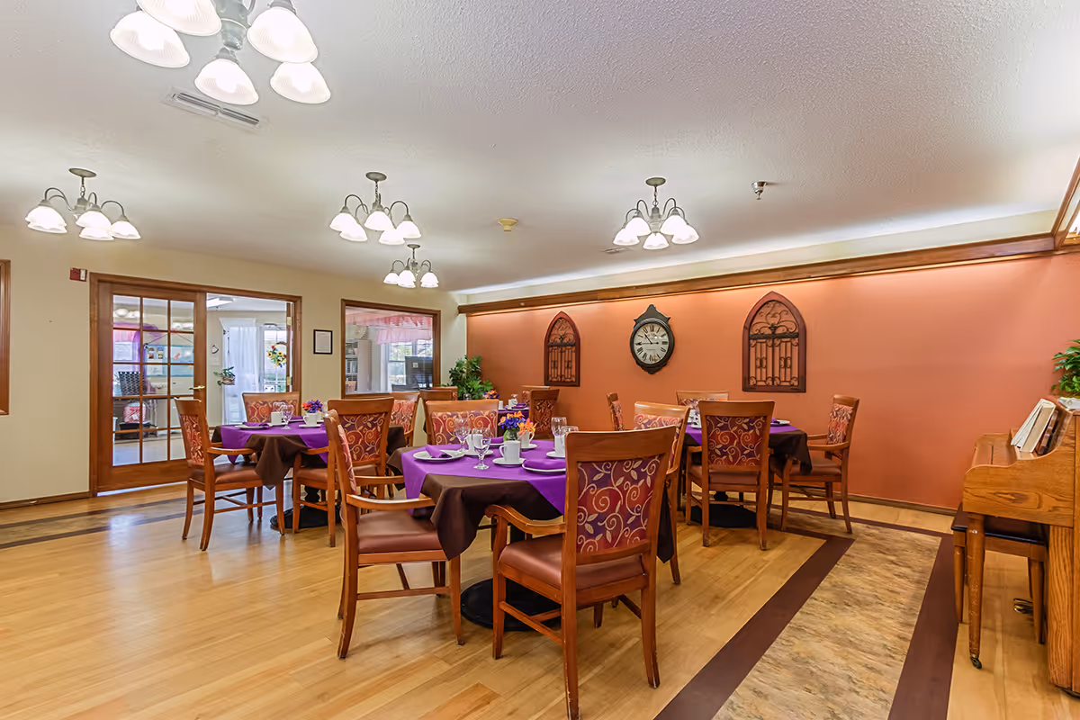 A bright dining room with several round tables set with purple tablecloths and wooden chairs against a warm orange accent wall.