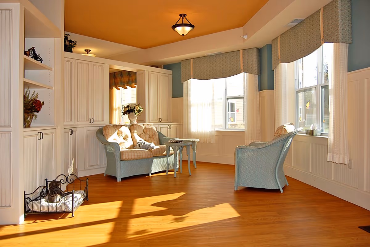 Sunlit common living room with wicker chairs and a loveseat by large windows and built-in cabinetry.