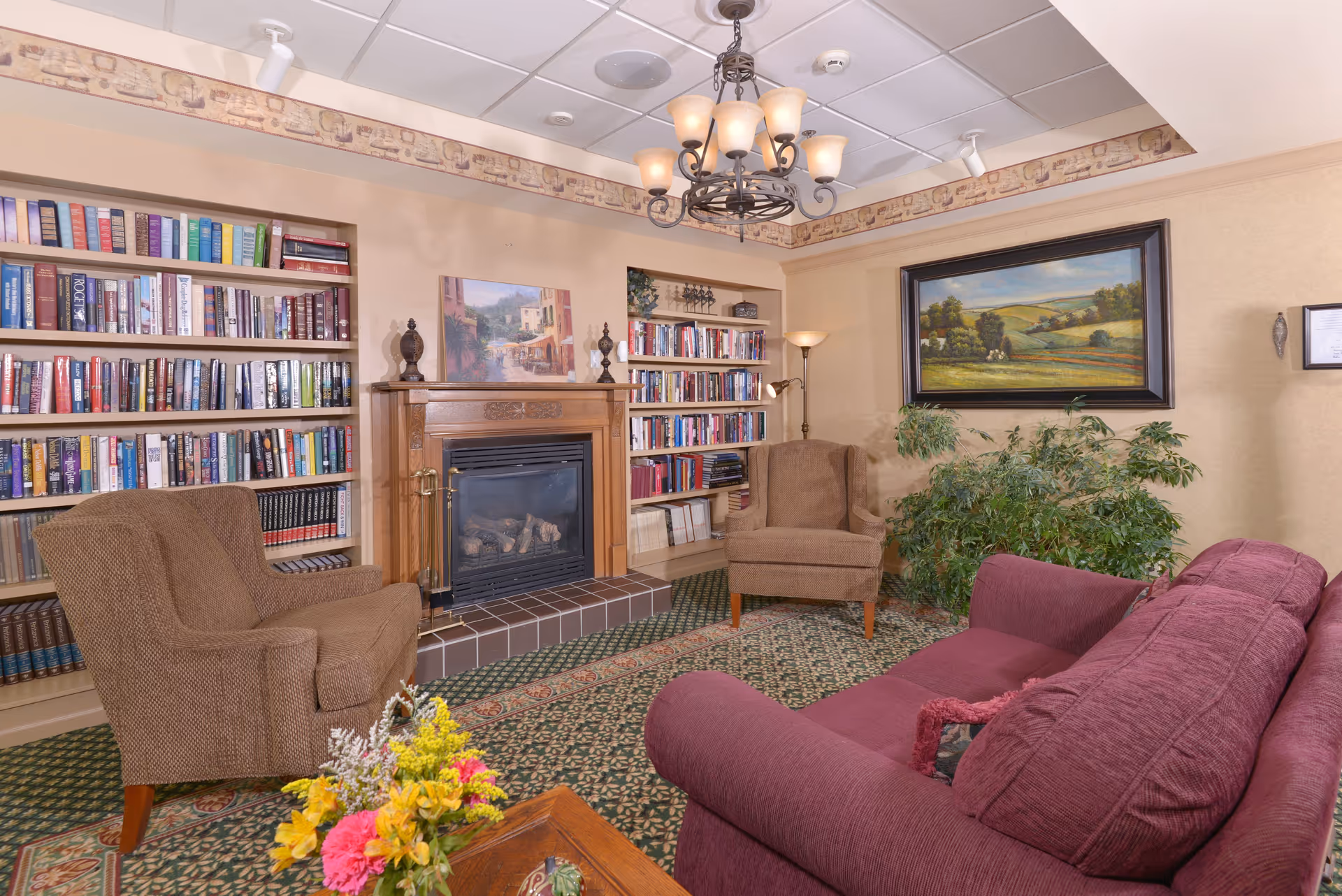 Cozy sitting room with bookshelves, a fireplace, armchairs, and a sofa decorated with artwork and plants.