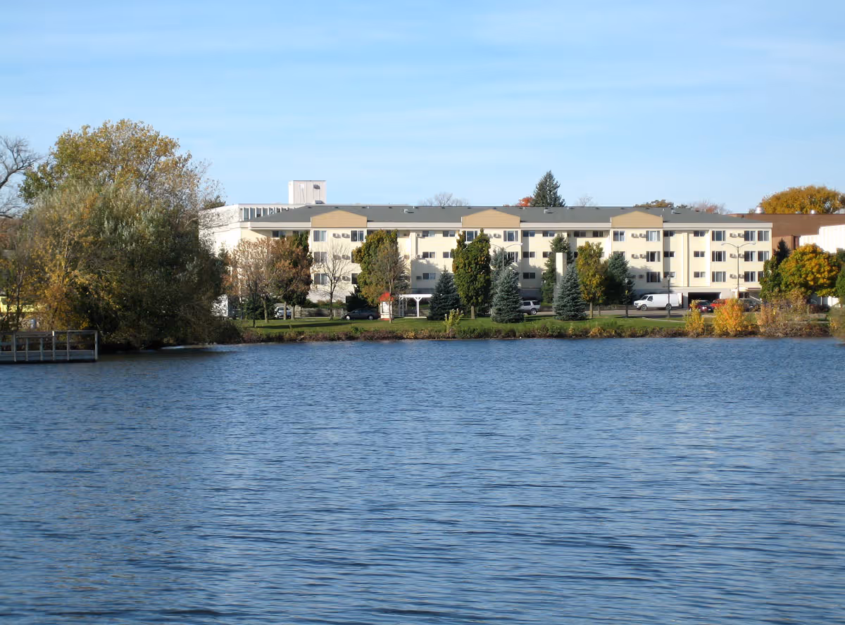 A large multi-story building viewed from across a body of water, surrounded by trees and greenery under a clear blue sky.