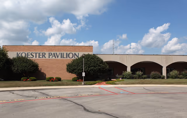 Exterior view of a single-story building named Koester Pavilion with a brick facade and arched entryways, surrounded by trimmed bushes and a clear blue sky with scattered clouds.