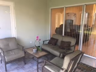 A small outdoor patio area with cushioned seating including a loveseat, two armchairs, and a coffee table with a potted orchid on it. The patio has tiled flooring and is adjacent to a sliding glass door with vertical blinds.