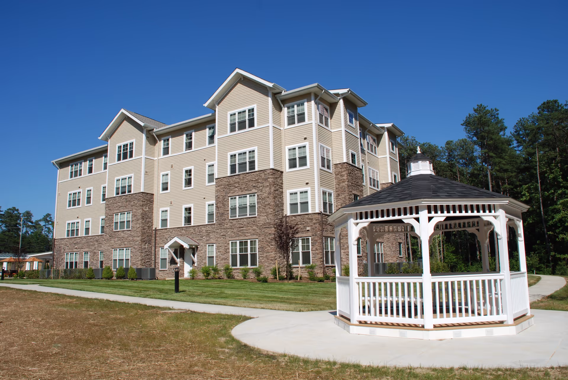 A multi-story senior living facility building with beige siding and stone accents under a clear blue sky. In the foreground, there is a white wooden gazebo on a concrete path surrounded by grass and trees.