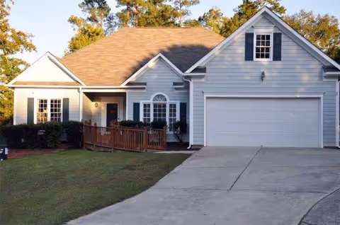Front exterior view of a single-story house with white siding, a brown shingled roof, a two-car garage, and a wooden ramp leading to the front door. The house is surrounded by a green lawn and trees in the background.