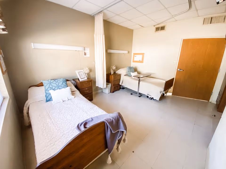 A tidy shared patient bedroom with two single beds, bedside tables, a privacy curtain between them, and a closed wooden door.