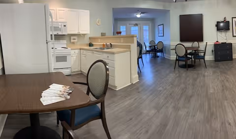 A spacious interior room featuring a kitchen area with white cabinets, a refrigerator, microwave, and stove. Adjacent to the kitchen are several round tables with chairs arranged for dining or socializing. The flooring is a light wood laminate, and the walls are painted light blue. In the background, there are double glass doors and framed artwork on the walls.