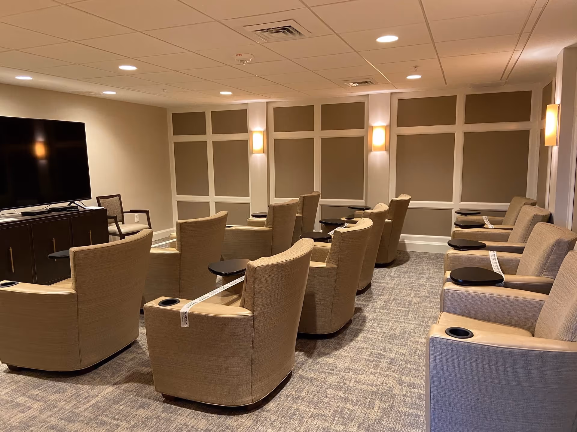 A cozy common area with multiple beige armchairs arranged in rows facing a large flat-screen TV mounted on a dark wooden cabinet. The room has soft lighting from wall sconces and a carpeted floor with a neutral pattern. Some chairs have white tape across them, possibly indicating social distancing measures.