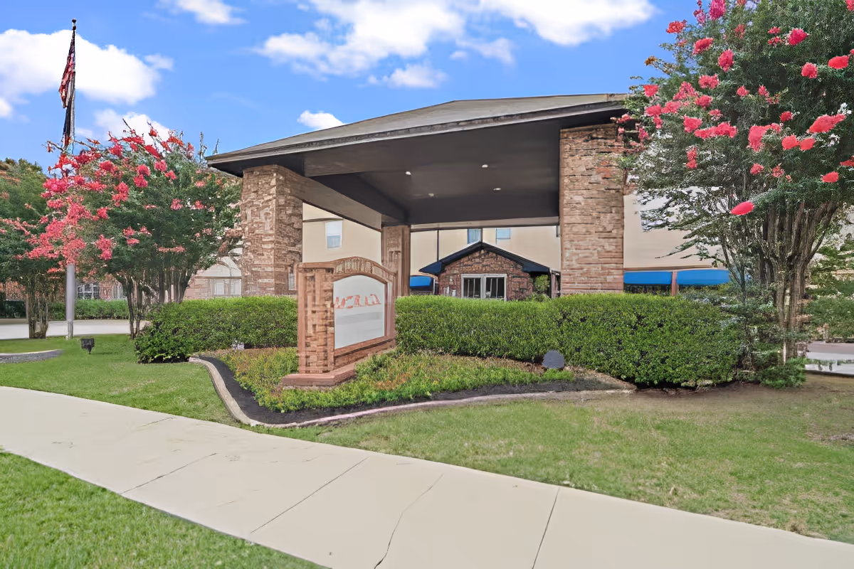 Entrance of Morada Meadow Lakes facility with a covered driveway supported by stone pillars, surrounded by green bushes and flowering trees under a blue sky with scattered clouds.