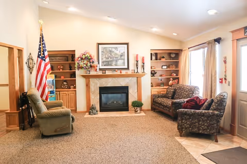 A cozy living room area with a fireplace in the center, flanked by built-in wooden shelves filled with decorative items. There is an American flag on the left side, a green recliner chair, and two patterned armchairs on the right side near a window with beige curtains. The room has carpeted flooring and a tiled entryway near the door on the right.