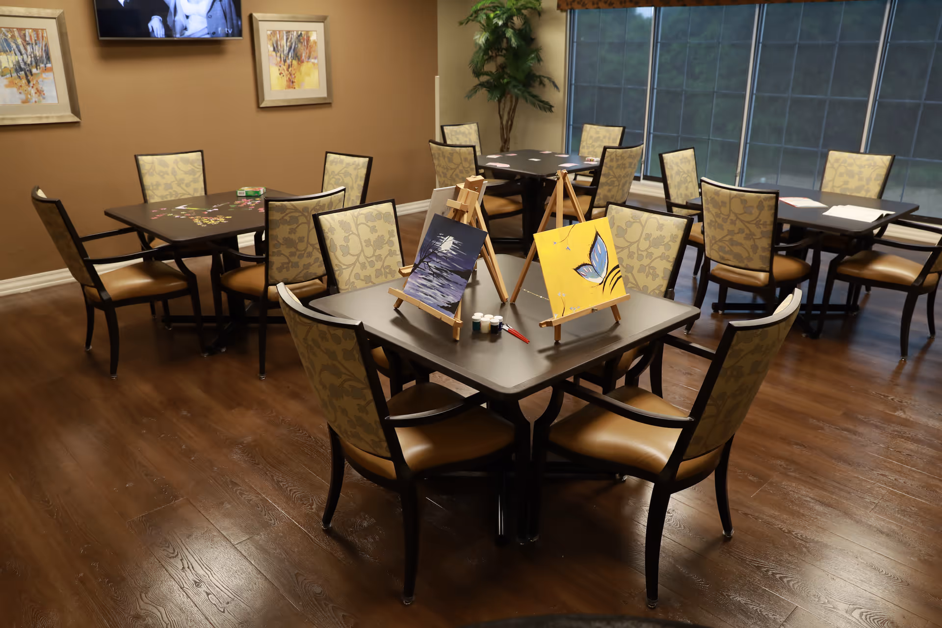 A well-lit room with several square tables surrounded by cushioned chairs with floral patterned backs. On the nearest table, there are two small easels holding paintings, along with paint pots and brushes, suggesting an art activity. The room has wooden flooring, large windows with blinds, and framed artwork on the walls. A television is mounted on the wall above one of the tables.