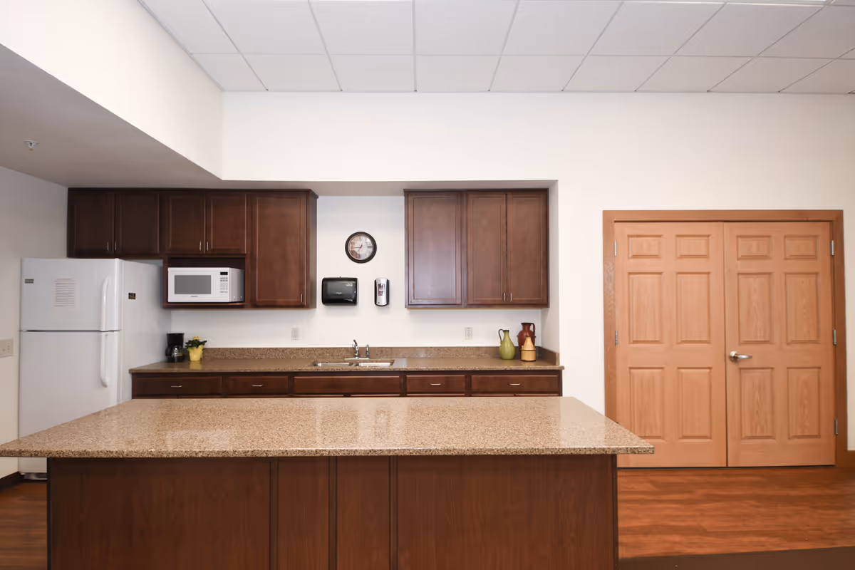 A clean kitchen area with dark wooden cabinets, a white refrigerator, a microwave, a coffee maker, a sink, and a large granite countertop island. There are decorative items on the counter and a clock, paper towel dispenser, and hand sanitizer mounted on the wall. Double wooden doors are visible on the right side.