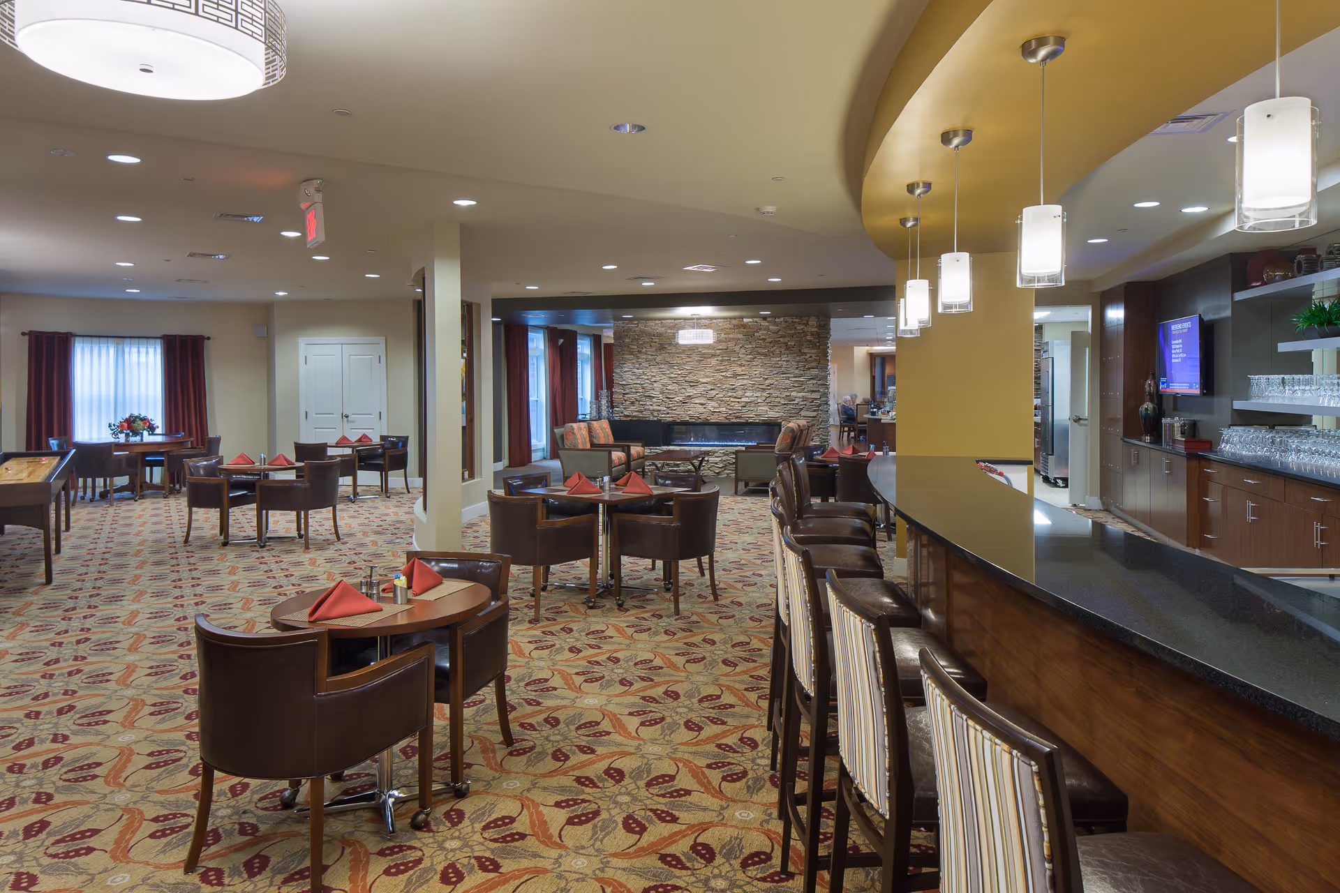 A spacious dining and lounge area in a senior living facility featuring multiple round tables with brown chairs and red napkins, a long bar counter with high chairs, pendant lights hanging from the ceiling, a stone accent wall with a modern fireplace, and large windows with curtains allowing natural light.