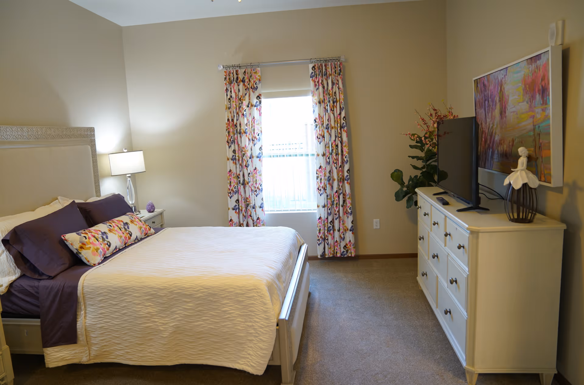 A neatly made bed with white and purple bedding in a bedroom. There is a bedside table with a lamp on the left side of the bed. A window with floral curtains is centered on the back wall. On the right side, there is a white dresser with a flat-screen TV, a decorative figurine, and a colorful painting hanging above it. A potted plant is placed next to the dresser.