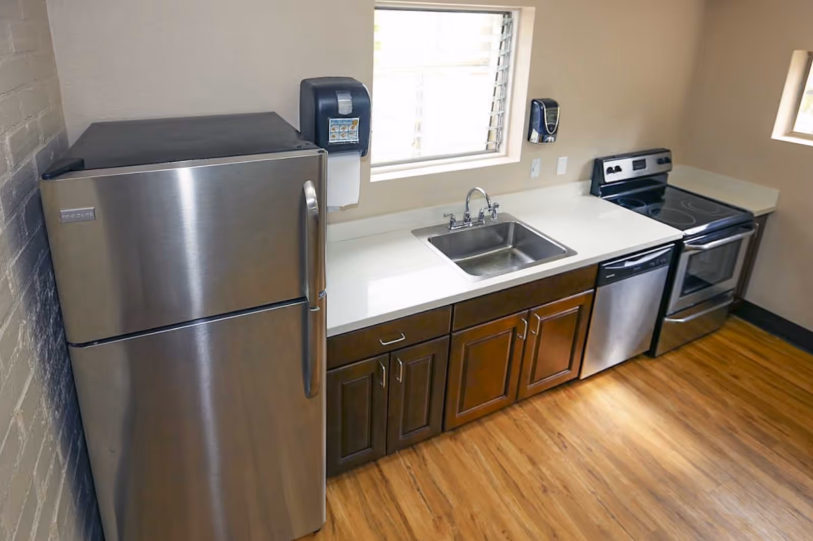 A kitchen area with stainless steel appliances including a refrigerator, dishwasher, and electric stove with oven. There is a white countertop with a sink and dark wooden cabinets below. The floor is wooden, and there are two windows letting in natural light.