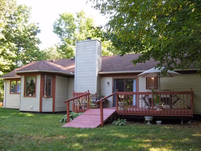 Single-story house with beige siding and a brown roof, featuring a wooden deck with a ramp and railing. The deck has outdoor furniture including a table with an umbrella and chairs. The house is surrounded by green grass and trees.