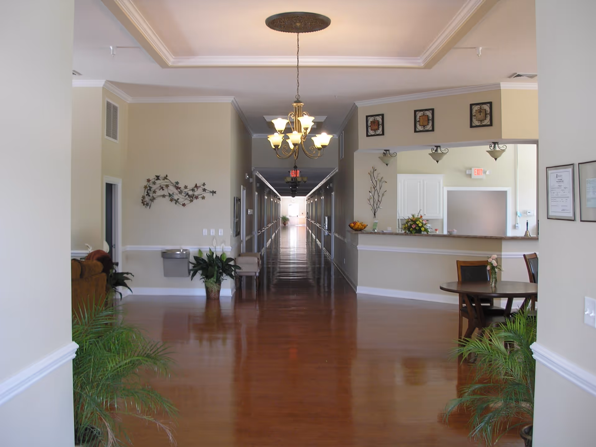 Reception area and long polished hallway of a senior living facility with chandeliers, seating, plants, and a front desk.