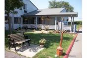 Outdoor view of a senior living facility with a white building featuring a covered porch. There is a bench on a concrete pad surrounded by grass and a small flower pot. The area is landscaped with hanging flower baskets and a red curb along the driveway.