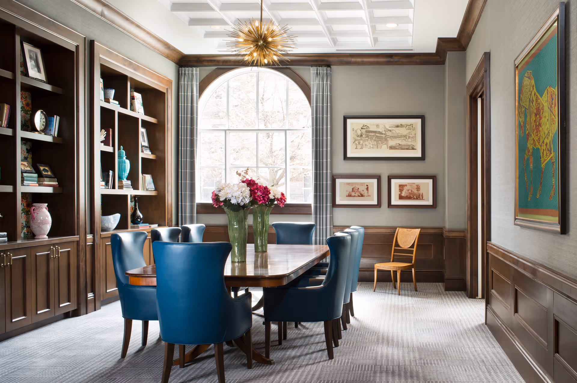 A formal dining room with a large wooden table surrounded by blue upholstered chairs. The room features built-in wooden bookshelves filled with decorative items and books, a large arched window with gray plaid curtains, and framed artwork on the walls. A modern gold chandelier hangs from the white coffered ceiling, and a vase with flowers is placed on the table.