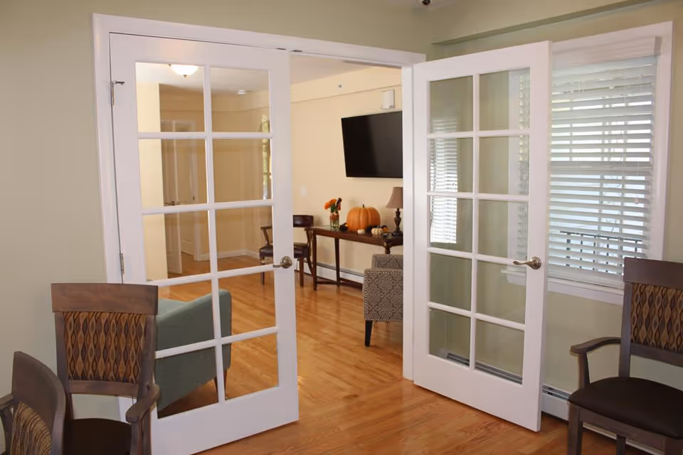 View through open white French doors into a bright living area with wood floors, chairs, a wall-mounted TV, and a table decorated with pumpkins.