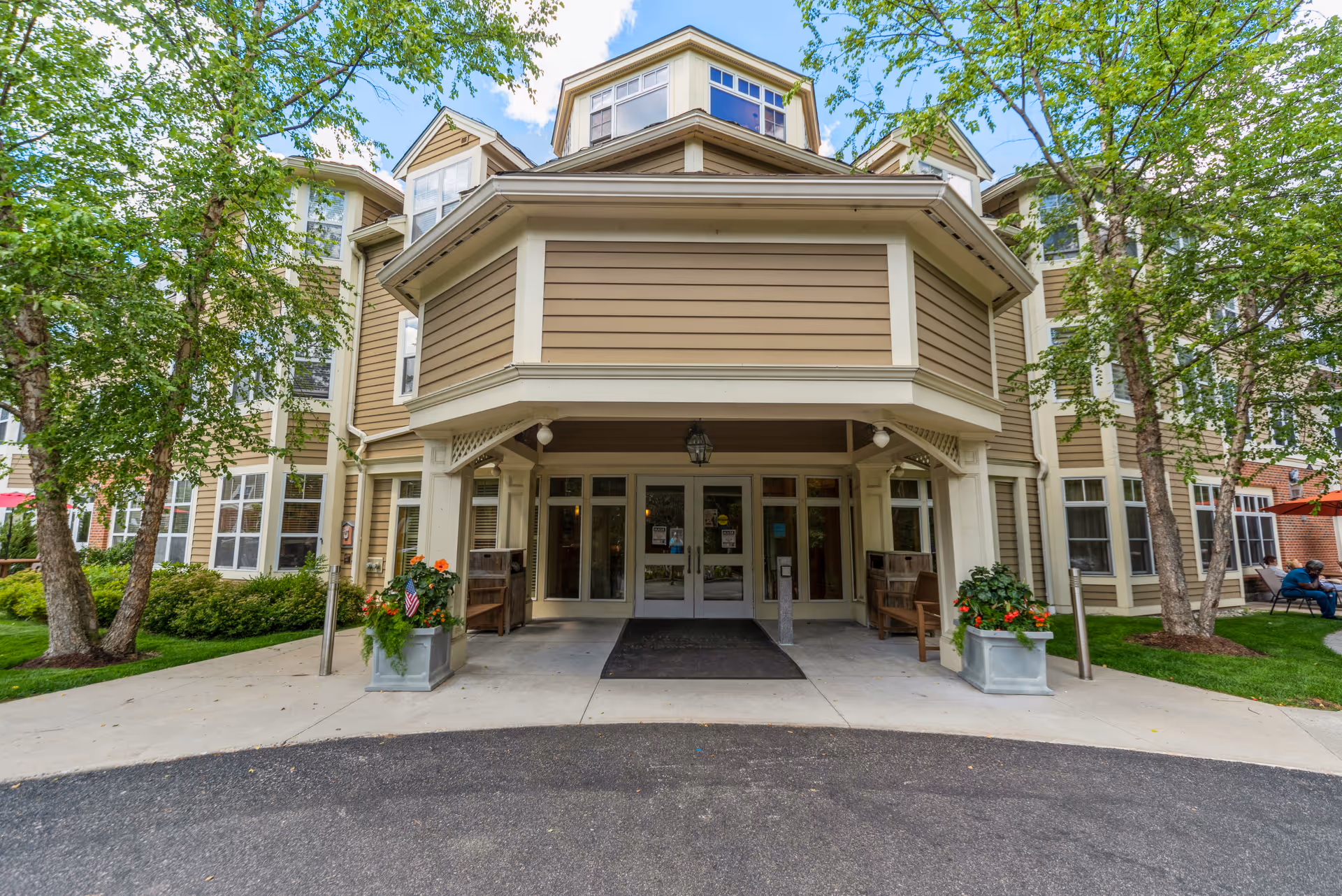 Front entrance of The Gables at Winchester senior living facility with a covered porch, double glass doors, potted plants with flowers on either side, and trees and greenery surrounding the building.