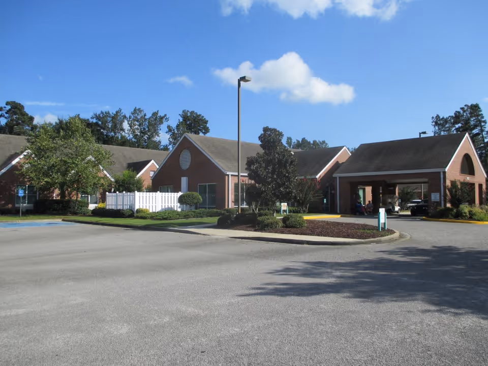 Exterior view of a single-story brick building with a covered entrance, surrounded by trees and landscaping under a blue sky with some clouds.