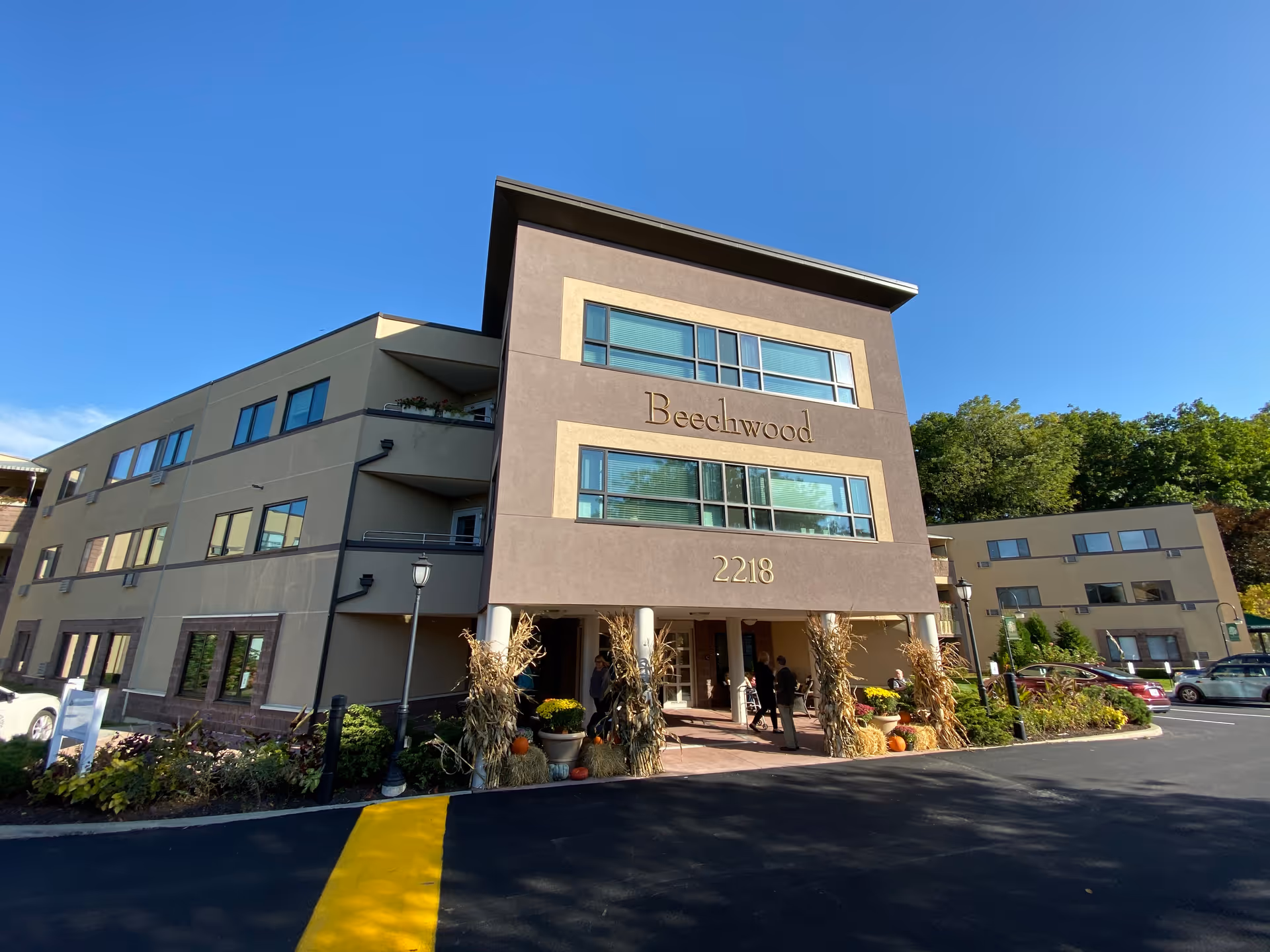 Front exterior of the Beechwood senior living building showing the main entrance, 'Beechwood' signage and address '2218', landscaping and parked cars under a clear blue sky.