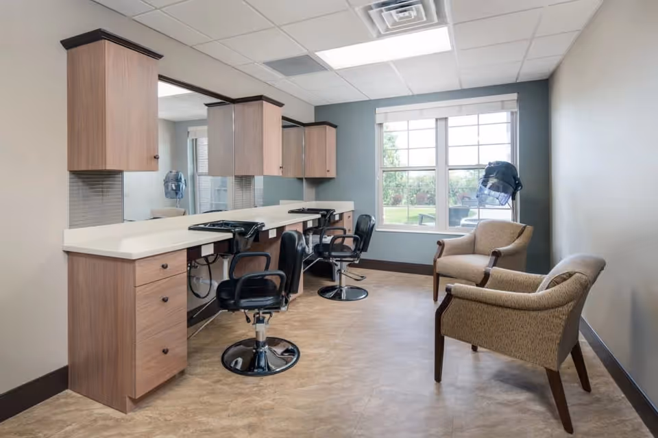 A bright salon area with two black styling chairs in front of a long counter with mirrors and wooden cabinets above. Two beige armchairs are placed near a large window that looks out onto a green outdoor space. A hair dryer hood is positioned near the window.