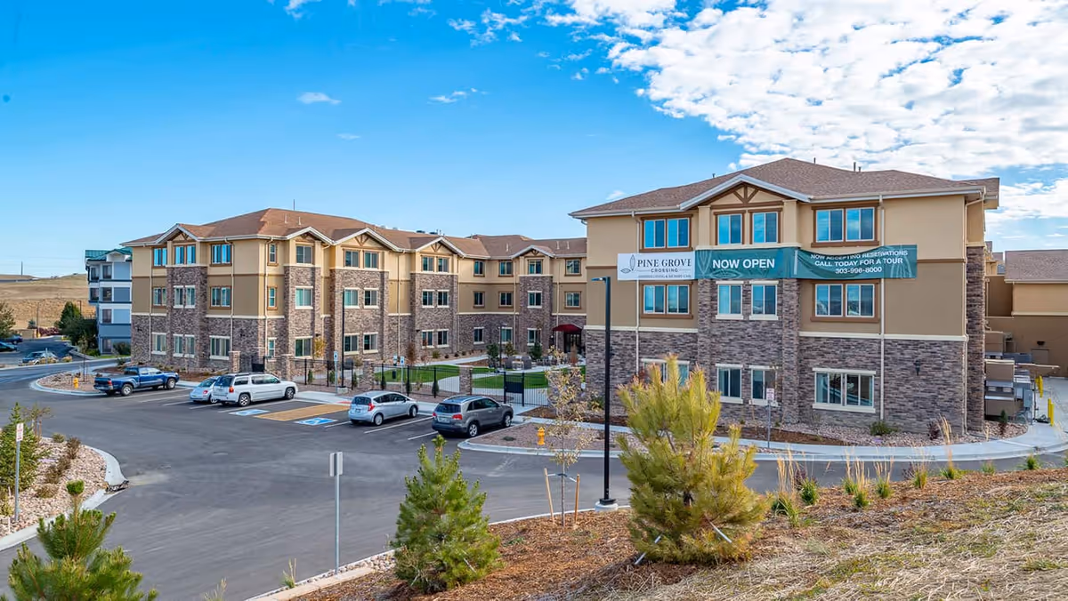 Exterior view of Pine Grove Crossing, a multi-story senior living facility with a stone and beige facade, several parked cars in the parking lot, and a clear blue sky with some clouds.