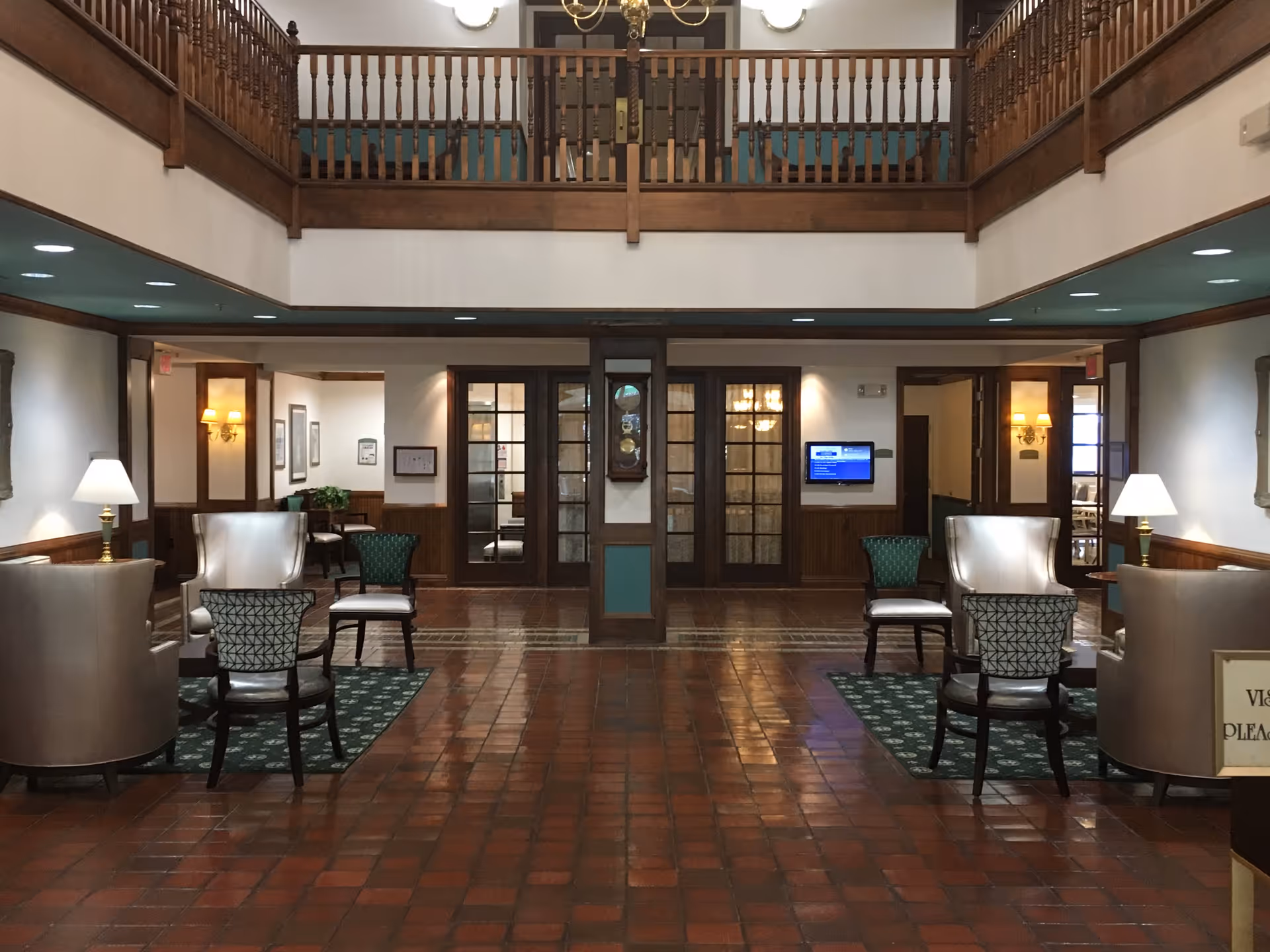 Interior view of a senior living facility lobby with a high ceiling and a second-floor balcony. The room features wooden paneling, tiled floor, several chairs arranged in small seating areas with lamps on side tables, and a wall clock mounted between two sets of glass doors.