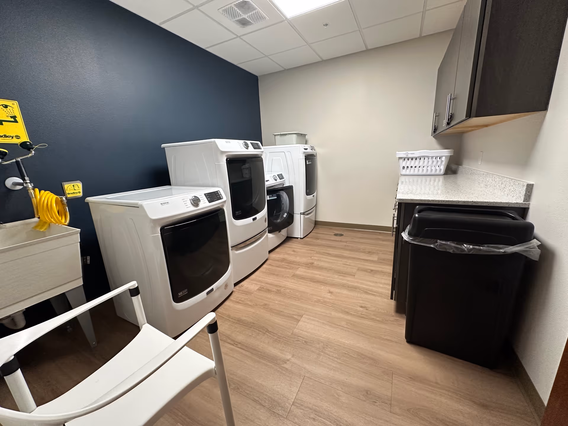 Laundry room with three white front-loading washing machines and dryers lined up against a wall. There is a white utility sink with a yellow coiled hose attached to the wall above it. A white chair is positioned in the foreground. On the right side, there is a countertop with cabinets above and below, and a black trash bin underneath the counter. The floor is wood-patterned, and the walls are painted in dark blue and white.
