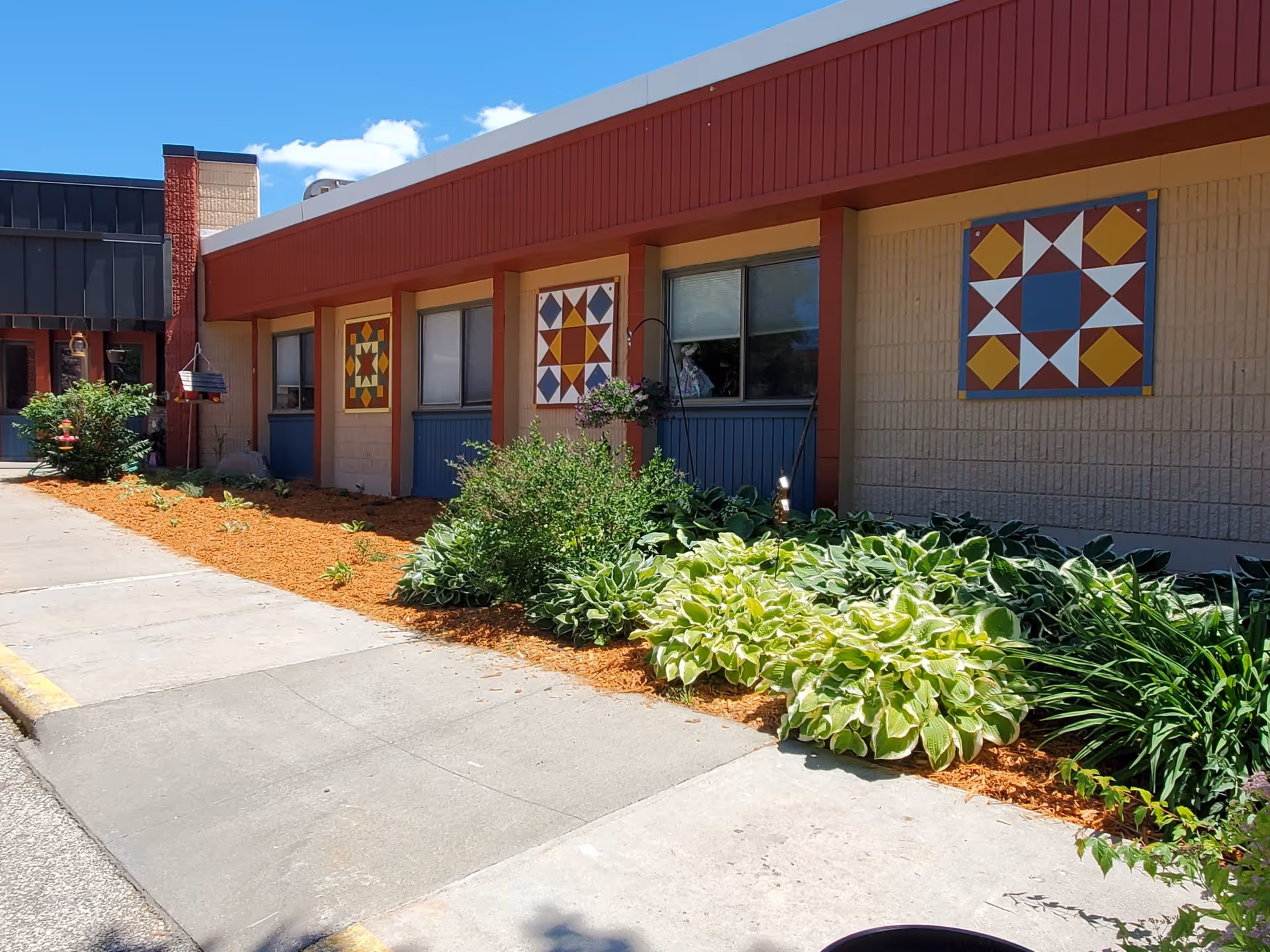 Exterior view of a senior living facility building with a garden bed along the sidewalk. The building has beige brick walls with red trim and three colorful geometric quilt patterns mounted on the wall. There are green shrubs and plants in the garden bed under a clear blue sky.