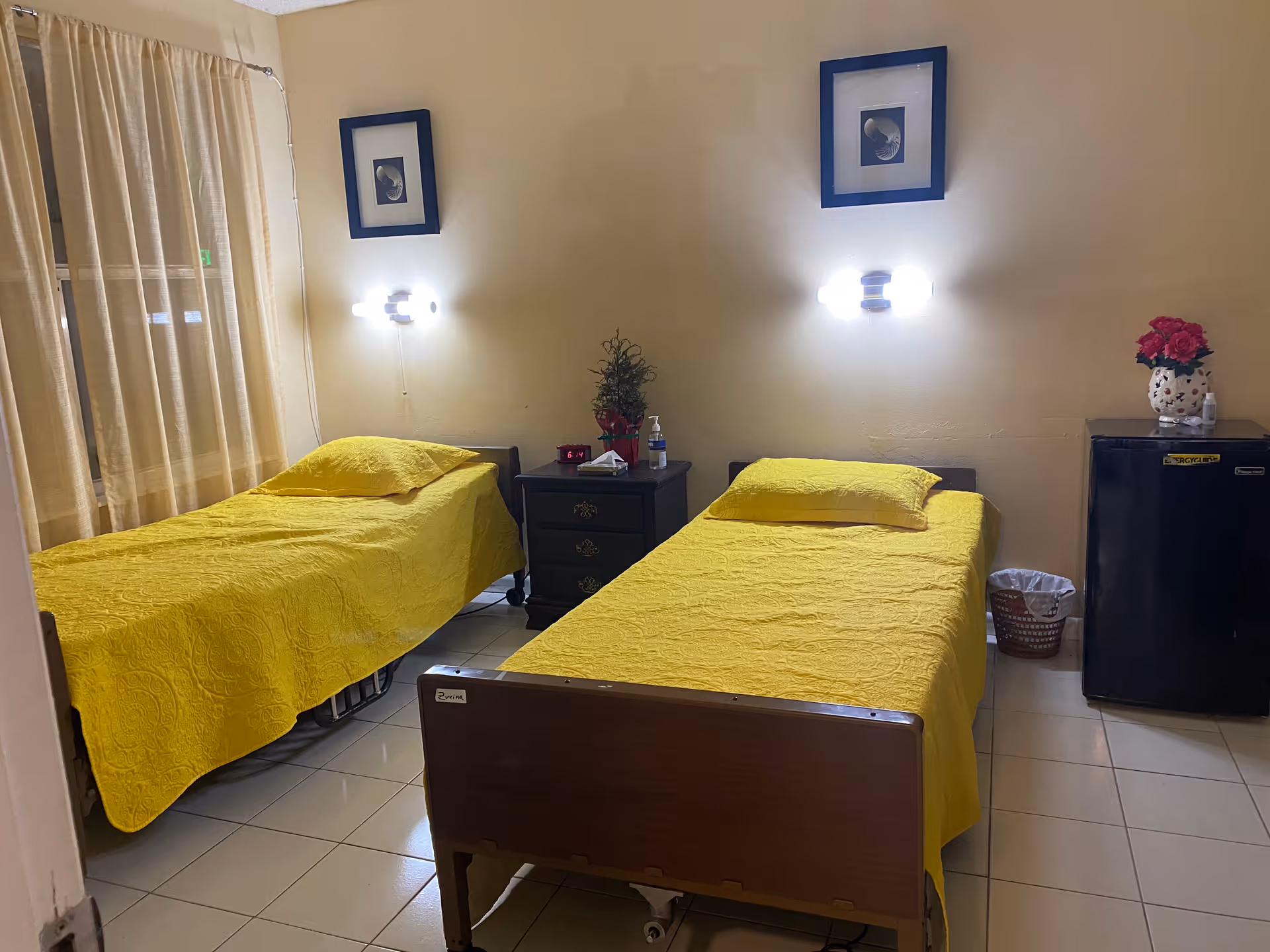 A bedroom with two single beds covered in yellow bedspreads. Between the beds is a small dark nightstand with a plant, a clock, and a bottle of hand sanitizer. Above each bed is a wall-mounted light and a framed picture. To the right, there is a small black refrigerator with a vase of red flowers on top and a wastebasket beside it. Light beige curtains cover the window on the left side of the room.