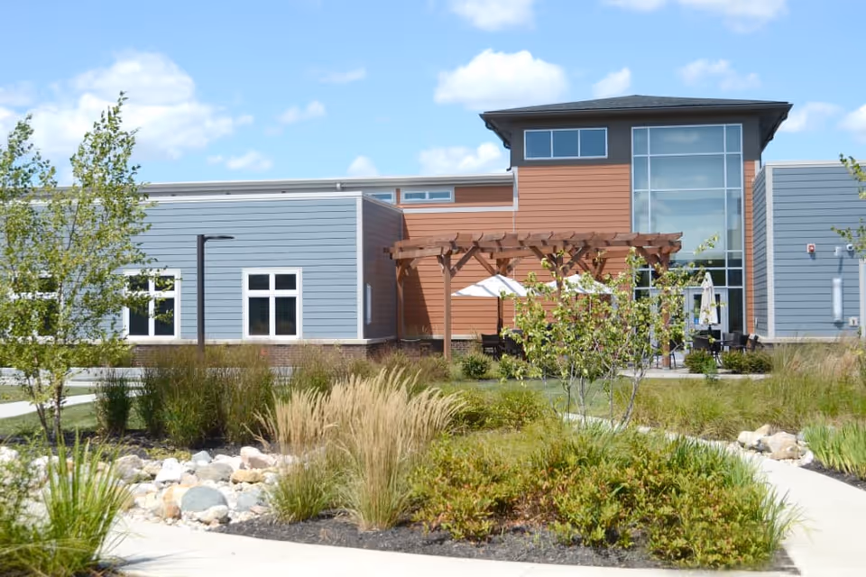 Modern senior living building exterior with landscaped garden, pergola-covered patio and large windows.